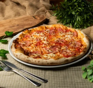 Na zdjęciu znajduje się pizza z serem i szynką na talerzu, otoczona roślinami i sztućcami na lnianym obrusie.