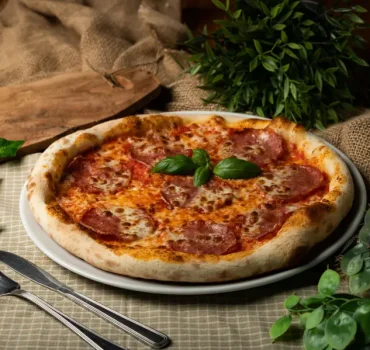 Na białym talerzu leży pizza z salami, serem i bazylią, umieszczona na tle rustykalnych dekoracji.