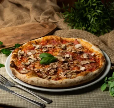 Na zdjęciu znajduje się pizza z szynką i pieczarkami, udekorowana liśćmi bazylii, podana na białym talerzu, obok sztućców.