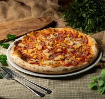 Na talerzu znajduje się pizza z szynką, ananasem i kukurydzą, podana na rustykalnym tle z zielonymi roślinami.