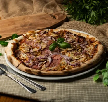 Na zdjęciu widoczna jest pizza z szynką, czerwonymi cebulami i świeżą bazylią, podana na białym talerzu, udekorowana zielonymi roślinami i sztućcami obok.