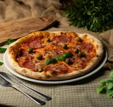Na zdjęciu widoczna jest pizza z salami, oliwkami i listkami bazylii, podana na białym talerzu.