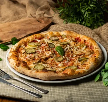 Na grafice znajduje się pizza z ogórkami kiszonymi, cebulą, mięsem i serem, podana na talerzu, otoczona zielonymi roślinami i sztućcami.