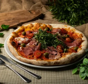Pizza z szynką, pomidorkami koktajlowymi, oliwkami i rukolą na rustykalnym tle.