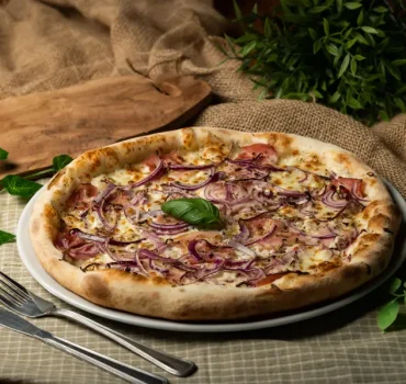Na zdjęciu widnieje pizza z szynką i czerwoną cebulą, udekorowana świeżą bazylią, podana na rustykalnym tle z zielonymi roślinami.