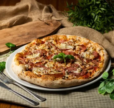 Na zdjęciu znajduje się apetyczna pizza z dodatkiem szynki, pieczarek i sera, podana na białym talerzu.
