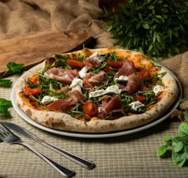 Na zdjęciu widać pizzę udekorowaną rukolą, pomidorkami koktajlowymi, szynką parmeńską i kremowym serem, podaną na talerzu, w otoczeniu zielonych roślin.