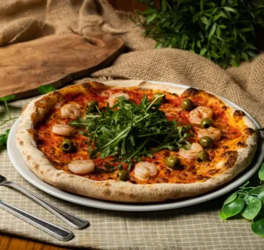 Pizza z krewetkami, oliwkami i rukolą na jasnym talerzu, otoczona zielonymi roślinami i sztućcami po bokach.