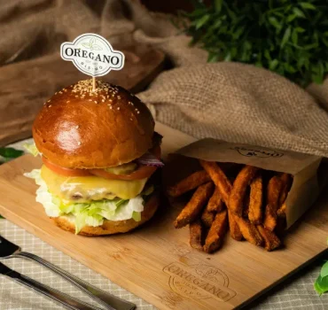 Na drewnianej desce znajduje się soczysty burger z warzywami i serem podany z frytkami z batatów w papierowym opakowaniu.