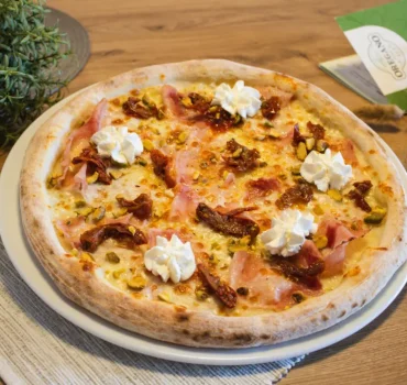 Na zdjęciu widać pizzę z plastrami szynki, suszonymi pomidorami, pistacjami i porcjami serka śmietankowego.
