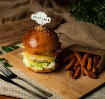 Na desce serwowany jest soczysty burger z warzywami i serem, obok frytki z batatów w papierowej torebce.