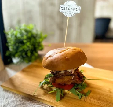 Na drewnianej desce znajduje się apetyczny burger z kotletem, zieleniną i pomidorem, wbity flagą z logo OREGANO Bistro.
