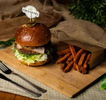 Apetyczny burger z warzywami, mięsem i serem podany na desce, obok frytki w brązowym papierowym opakowaniu.